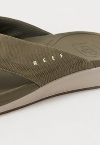 Žalios šlepetės su tekstūruotu viršumi, lygiu padu ir spausdintu "REEF" logotipu ant dirželio. Lengvos ir skirtos kasdieniam dėvėjimui.