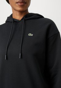 Svart hettegenser laget av mykt materiale med kengurulomme. Har snorhette og et lite brodert grønt krokodille-logo på brystet.