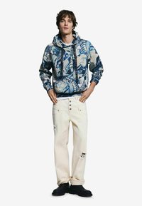 Modrá a krémová mikina s kapucňou s abstraktnými vzormi, doplnená krémovými džínsami s ošúchanými detailmi a čiernou obuvou.