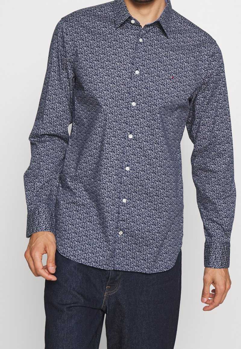 Homme portant une chemise bleu marine à manches longues avec de petits motifs floraux blancs et un jean bleu foncé, debout avec les mains relâchées le long du corps.