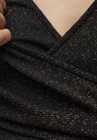 Μαύρο ύφασμα με λεπτομέρειες χρυσής λάμψης κρατημένο κοντά στον λαιμό σχηματίζοντας σχήμα V σε ανοιχτόχρωμο δέρμα.