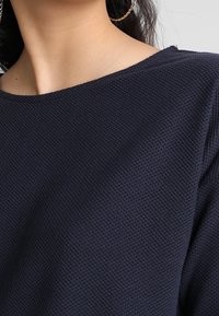 Marineblå stickad topp med en texturerad, våffelliknande mönster. Har en båt-krage och en avslappnad passform. Nära vy av tyget och halsringningen.