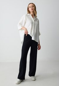 Camicia oversize bianca in lino con maniche corte, abbinata a pantaloni ampi a coste neri e sneakers bianche. Silhouette pulite e rilassate.