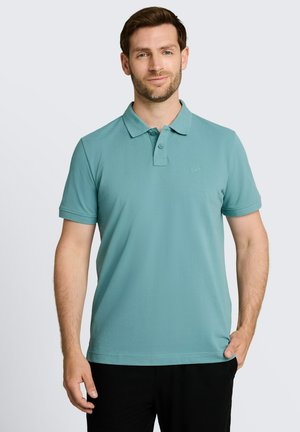 Man draagt een lichtblauw poloshirt met korte mouwen en een zwarte broek, staand met één hand in de zak tegen een effen achtergrond.