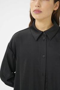 Černá košile na knoflíky s hladkou, lesklou texturou. Má klasický límec a uvolněný střih s spuštěnými rameny.