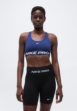 Športovo zdatná žena stojí čelom dopredu, oblečená v tmavomodrej športovej podprsenke Nike Pro a čiernych cyklistických šortkách Nike Pro, na neutrálom pozadí.