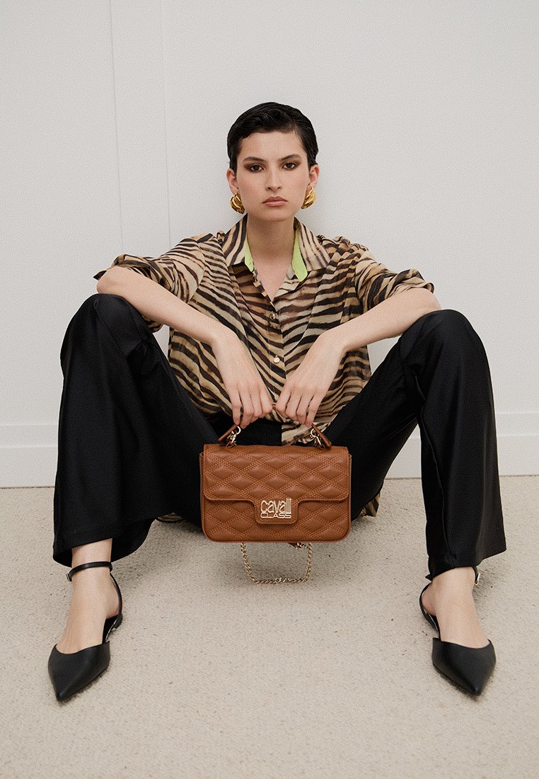 Borsa a tracolla marrone trapuntata con hardware dorato, abbinata a una camicia a righe marrone e beige e pantaloni neri a gamba larga. Presenta scarpe nere a punta.