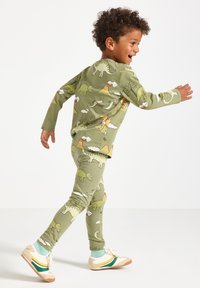 Un ragazzo sorridente che cammina, indossando un pigiama con stampa di dinosauri verdi e sneaker bianche con strisce verdi e gialle.