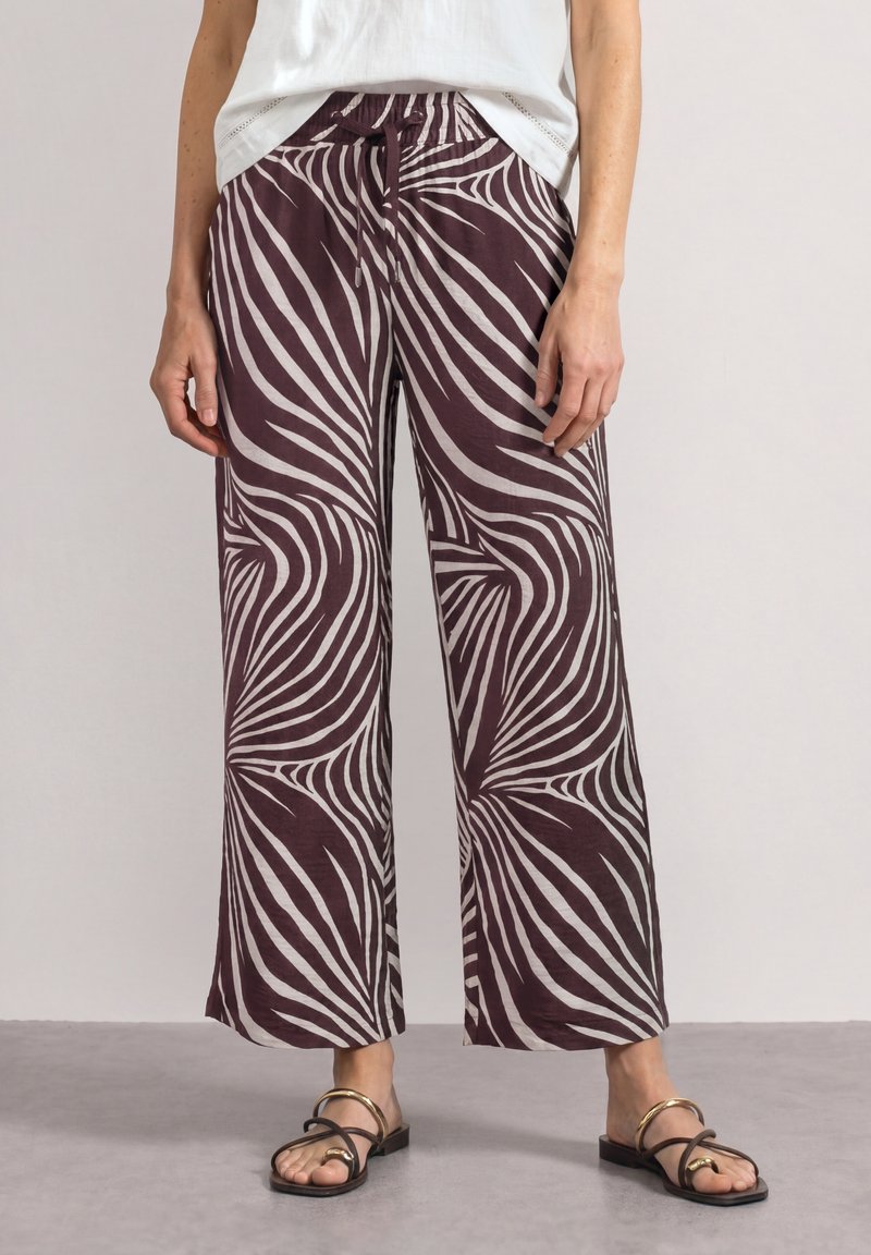 Femme portant un pantalon à jambes larges avec des rayures abstraites violettes et blanches, associé à des sandales plates noires à brides dorées.