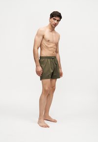 Homme torse nu debout pieds nus portant un short de bain vert olive avec cordon sur fond blanc uni.