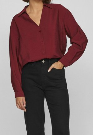 Femme portant une chemise bordeaux à manches longues boutonnée, rentrée dans un pantalon noir taille haute, une main dans la poche.