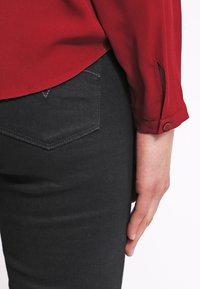 Rote Bluse mit abgerundetem Saum und geknöpften Manschetten, kombiniert mit schwarzen Jeans, die eine rechteckige Gesäßtasche und strukturierten Stoff aufweisen.