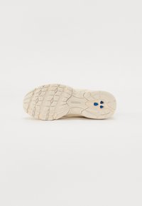 Semelle d'une basket beige New Balance avec une surface texturée antidérapante et trois petits détails bleus près du talon sur fond blanc.