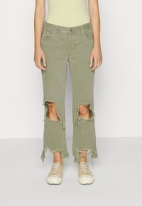 Jeans rasgados de un verde olivo claro con dobladillos deshilachados y grandes cortes en las rodillas, combinados con zapatillas beige. Material de algodón, ajuste casual.