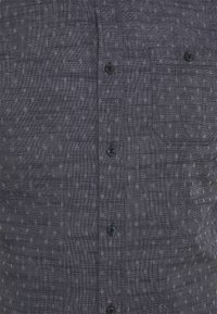 Camisa abotonada en tela gris oscuro, con una sutil textura estampada, dos botones delanteros y un bolsillo en el pecho.