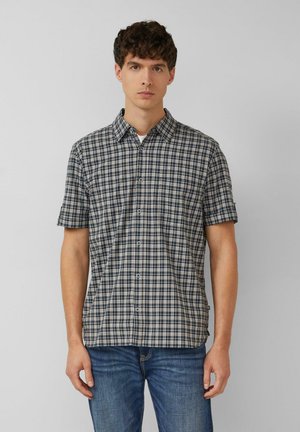 Mężczyzna ubrany w koszulę na guziki z krótkim rękawem w czarno-biało-niebieską kratę oraz niebieskie dżinsy, stojący na tle jednolitego szarego tła.