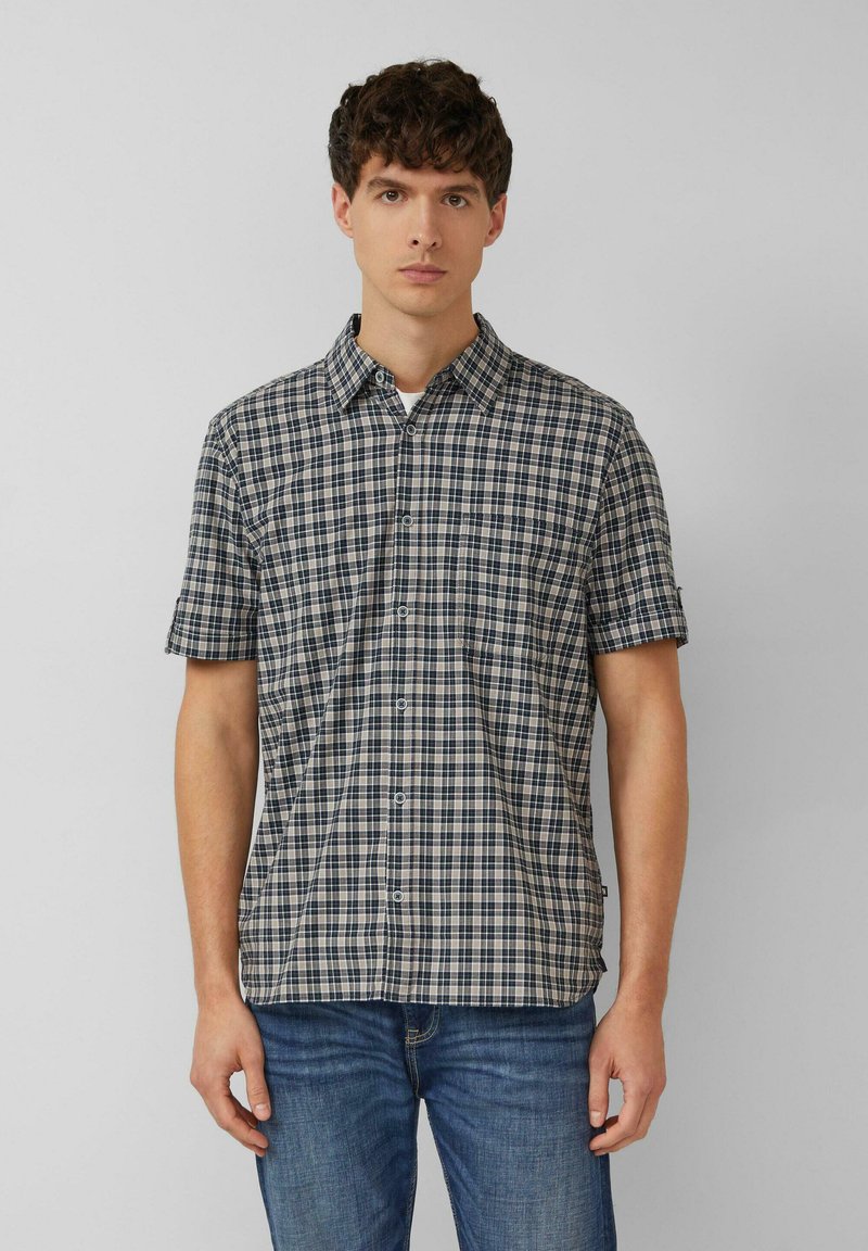 Mann trägt kurzärmliges Hemd mit schwarz-weiß-blauem Karomuster und blaue Jeans, steht vor einfachem grauem Hintergrund.