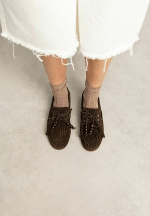 Feet wearing beige ribbed socks and dark brown suede moccasins with fringe and beaded laces, standing on a light gray floor.