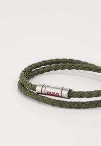 Bracciale intrecciato in pelle verde oliva con chiusura metallica argentata contrassegnata dal testo rosso "HUGO", texture liscia e design regolabile.