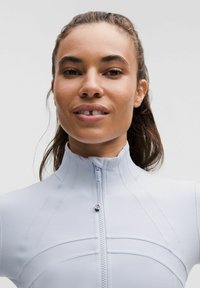 Žena s pevným drdolem, oblečená v světle modré sportovní mikině na zip, jemně se usmívající na neutrálním pozadí.