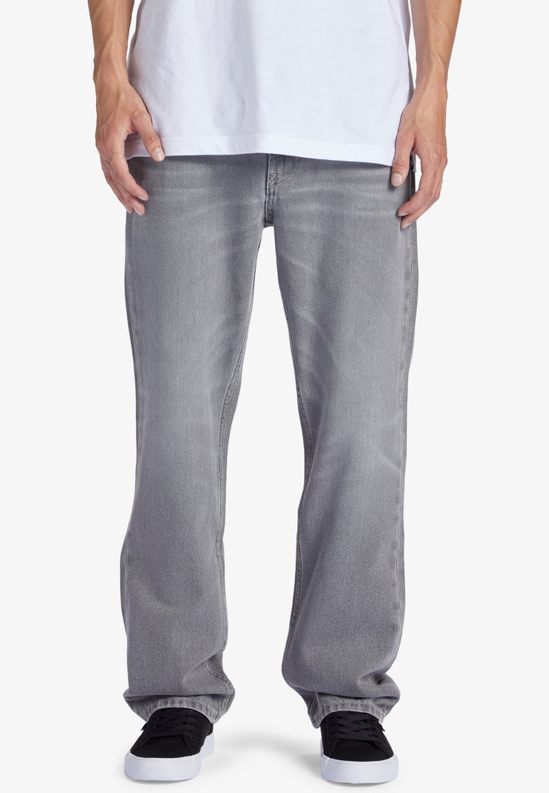 Personne portant un jean gris ample, des baskets noires avec des semelles blanches et un T-shirt blanc, debout devant un fond blanc.