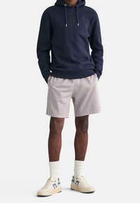 Granatowa bluza z kapturem z wytłoczonym logo, jasnoszare szorty i kremowe sneakersy z niebieskimi akcentami. Model stoi z rękami w kieszeniach.