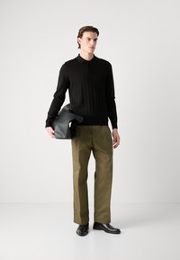 Cămașă polo neagră cu mânecă lungă și striații, pantaloni verzi măslin și pantofi negri. Modelul ține o geantă din piele neagră. Fundal neutru.