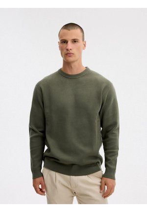 Un hombre luce un jersey liso de cuello redondo en color verde oliva y unos pantalones beige sobre un fondo blanco de estudio.