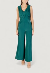 Modelo vistiendo un mono teal con un escote profundo en V y pantalones de pierna ancha, combinado con tacones nude frente a un fondo claro.