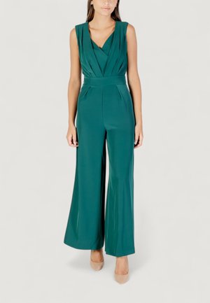 Modella che indossa una tuta teal con scollatura a V profonda e pantaloni a gamba larga, abbinata a tacchi nudi su uno sfondo chiaro.