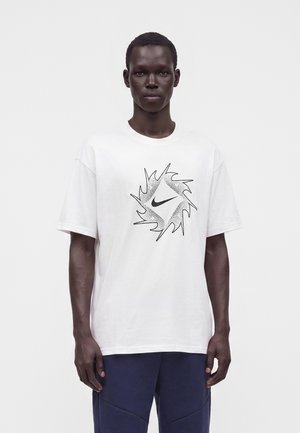 Άνδρας που φοράει λευκό T-shirt με μαύρο λογότυπο Nike μέσα σε σχεδιασμό με οδοντωτό κυκλικό σχήμα, συνδυασμένο με σκούρο μπλε παντελόνι, στέκεται σε λευκό φόντο.