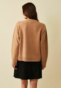 Pitkät ruskeat hiukset omaava nainen pukeutuneena löysään kamelinväriseen neulepaitaan ja mustaan, ohuella raidoituksella varustettuun hameeseen, kuvattu takaapäin yksinkertaisen taustan edessä.