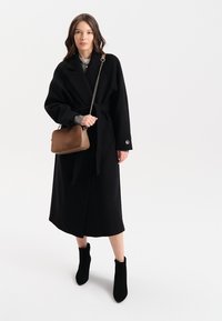 Czarny wełniany płaszcz z szerokimi rękawami, z paskiem w talii i detalem z guzikiem. W połączeniu z czarnymi botkami za kostkę i brązową skórzaną torebką na ramię.