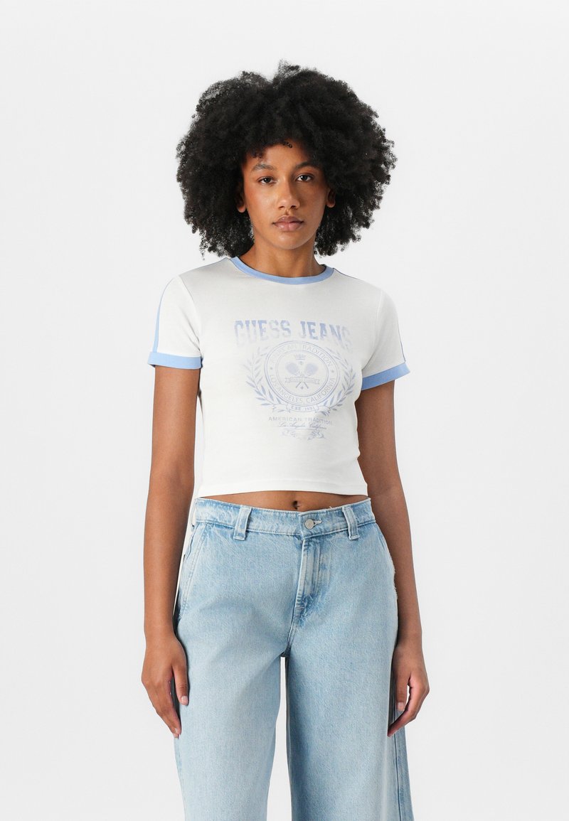 Valge lühikeste varrukatega särk sinise krae ja varruka aktsentidega; esiküljel on helesinine "GUESS JEANS" graafika. Kombineeritud helesiniste teksadega.