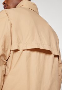 Veste beige clair en tissu texturé, avec un col rabattu et un détail de rabat à l'arrière. Design simple et moderne avec des coutures visibles.
