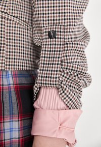 Manche en tissu à motif pied-de-poule brun et noir avec un bouton noir, superposée à une manche texturée rose clair avec un détail de nœud.