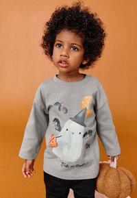 Szara koszula z długim rękawem z grafiką ducha w czapce czarownicy, nietoperzy i dyni. Mieszanka bawełny, noszona z ciemnymi spodniami i trzymająca pluszową dynię.