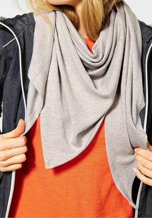 Écharpe gris clair en tissu doux, drapée lâchement autour du cou, superposée sur un haut orange, avec des mains tenant les bords de l'écharpe.