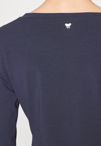 Maglietta a maniche lunghe blu navy in morbido cotone. Caratterizzata da un ampio scollo e un piccolo logo bianco sul retro, vicino al collo.