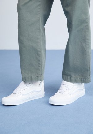 Piernas con pantalones verdes y zapatillas blancas bajas, de pie sobre un suelo azul claro contra un fondo blanco.