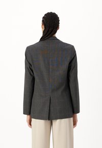 Grå rutete blazer med lange ermer, strukturerte skuldre og en enkelt bakre ventil, laget av teksturert stoff. Bukse er lys beige.