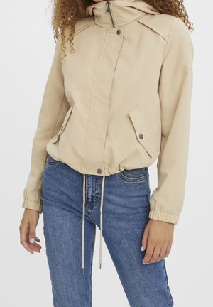 Femme portant une veste beige zippée avec poches à pressions et cordon à la taille, associée à un jean bleu, devant un fond uni.