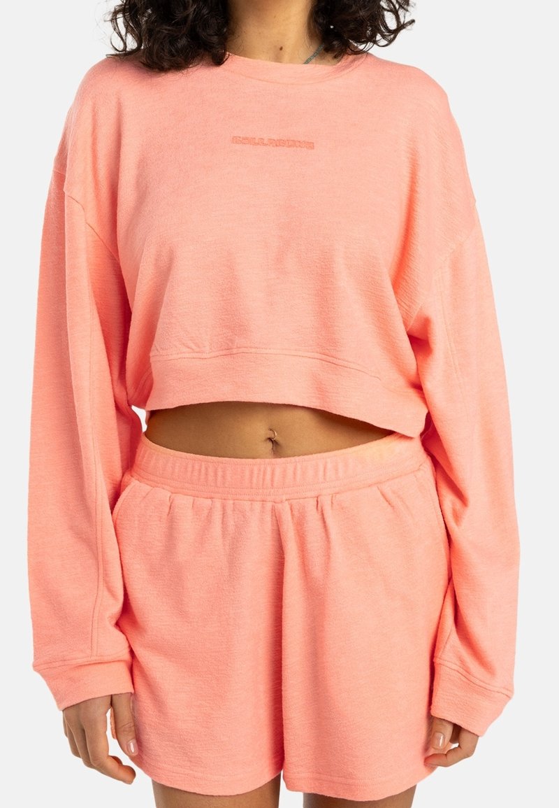 Sweat-shirt court rose clair à manches longues, avec une texture douce et un logo brodé. Assorti à un short taille haute assorti.