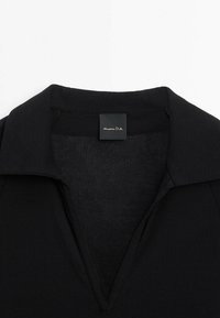 Chemise en maille côtelée noire avec col et ouverture en V, avec une petite étiquette noire portant le texte blanc « Massimo Dutti » à l'intérieur du col.
