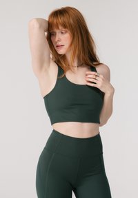 Rødhåret kvinne poserer iført mørkegrønn sports-BH og tights med høy midje mot en enkel lys bakgrunn.