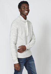 Hellgrauer Strickpullover mit Kapuze und weißen Kordeln. Gerippte Bündchen und Saum, mit texturiertem Muster, kombiniert mit dunkelblauen Jeans.