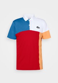 Koszulka polo w blokach kolorów niebieskiego, białego, czerwonego i pomarańczowego. Materiał jest gładki, posiada kołnierzyk i plisę z trzema guzikami oraz logo.