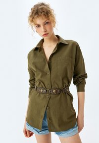Camisa de botones verde oliva con cuello, combinada con un cinturón marrón con tachuelas de metal, usada sobre pantalones cortos de mezclilla claros.