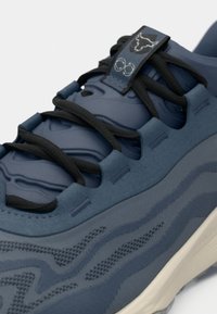 Chaussure de sport bleue avec une tige texturée à motif ondulé, lacets noirs plats et une semelle de couleur crème contrastante.