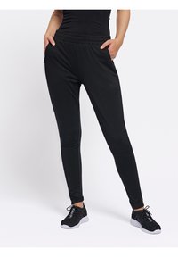 Osoba ubrana w czarne spodnie sportowe i czarne buty sportowe z białymi podeszwami, stoi z rękami w kieszeniach na białym tle.